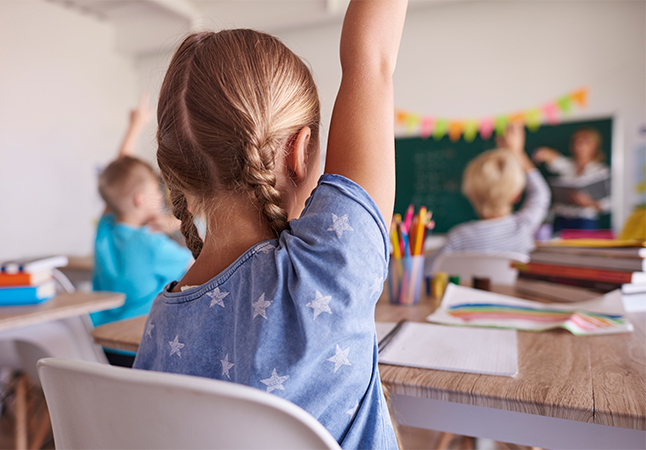 Foto di una bambina di spalle che alza una mano in classe
